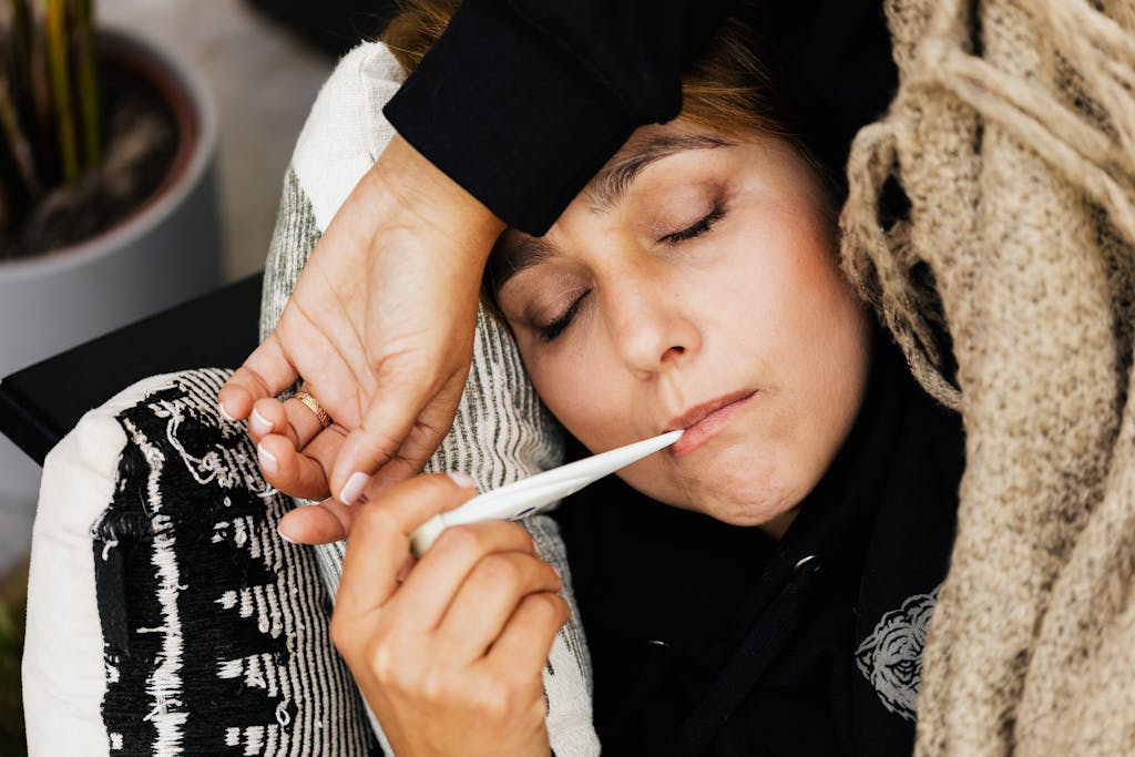 A woman resting on a couch using a thermometer to check her temperature, wrapped in a cozy blanket.