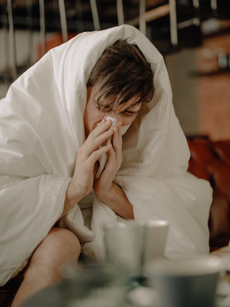 A young man wrapped in a blanket sneezes, indicating illness or cold symptoms.