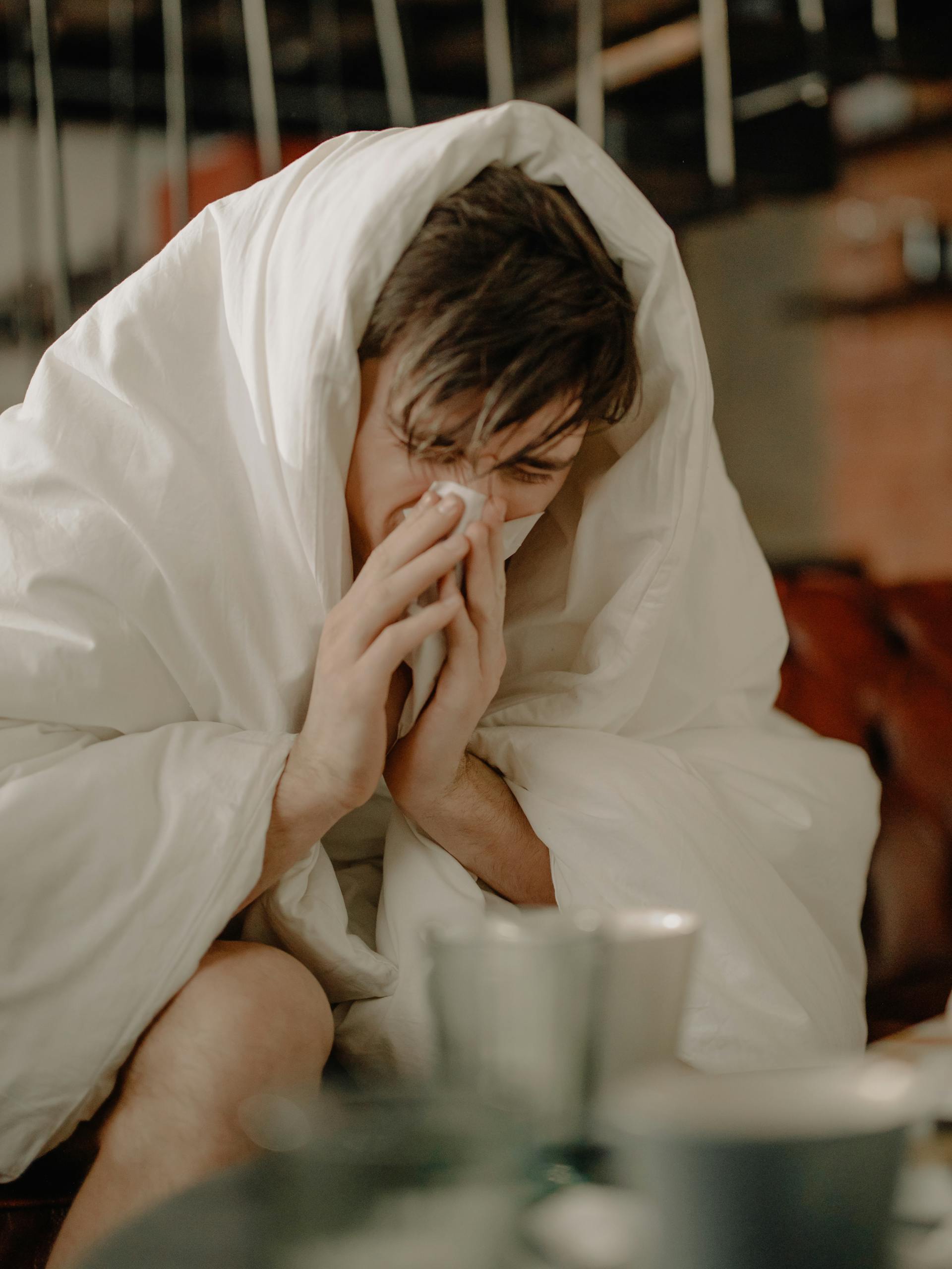 A young man wrapped in a blanket sneezes, indicating illness or cold symptoms.