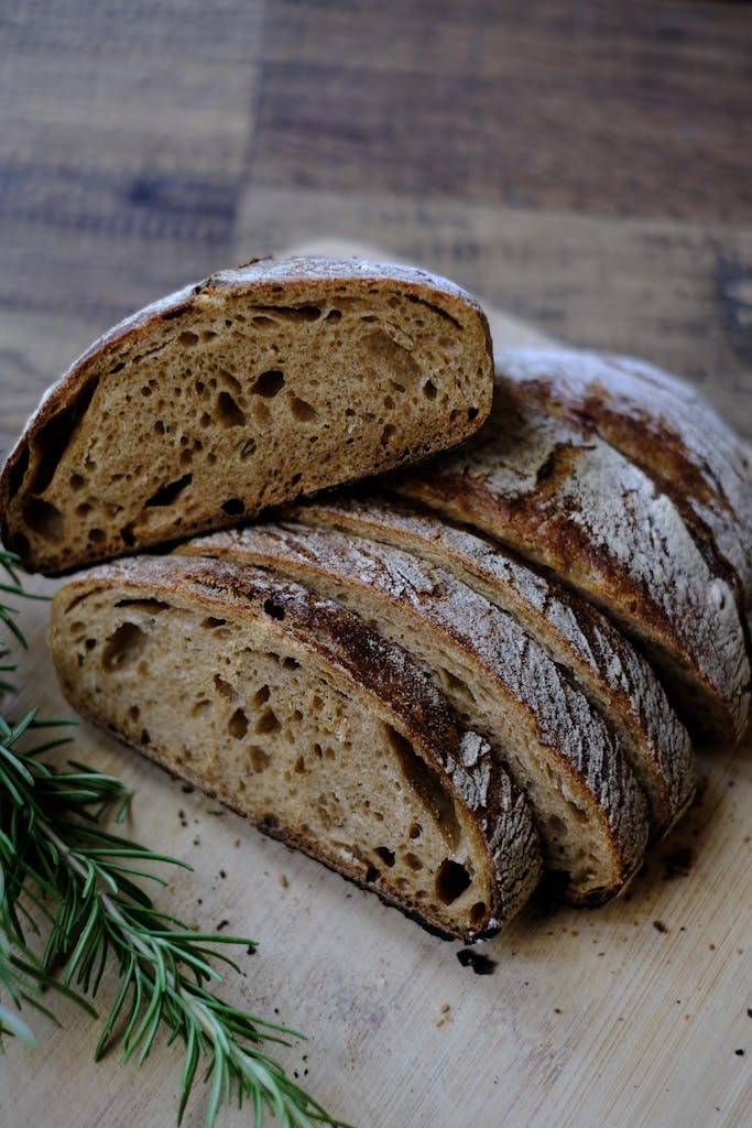 Close-up of a sliced, crusty sourdough loaf with fresh rosemary on a wooden surface.