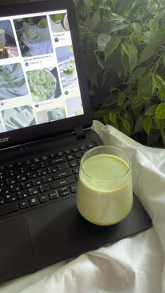 Glass of green milk drink on laptop keyboard amidst lush indoor plants.