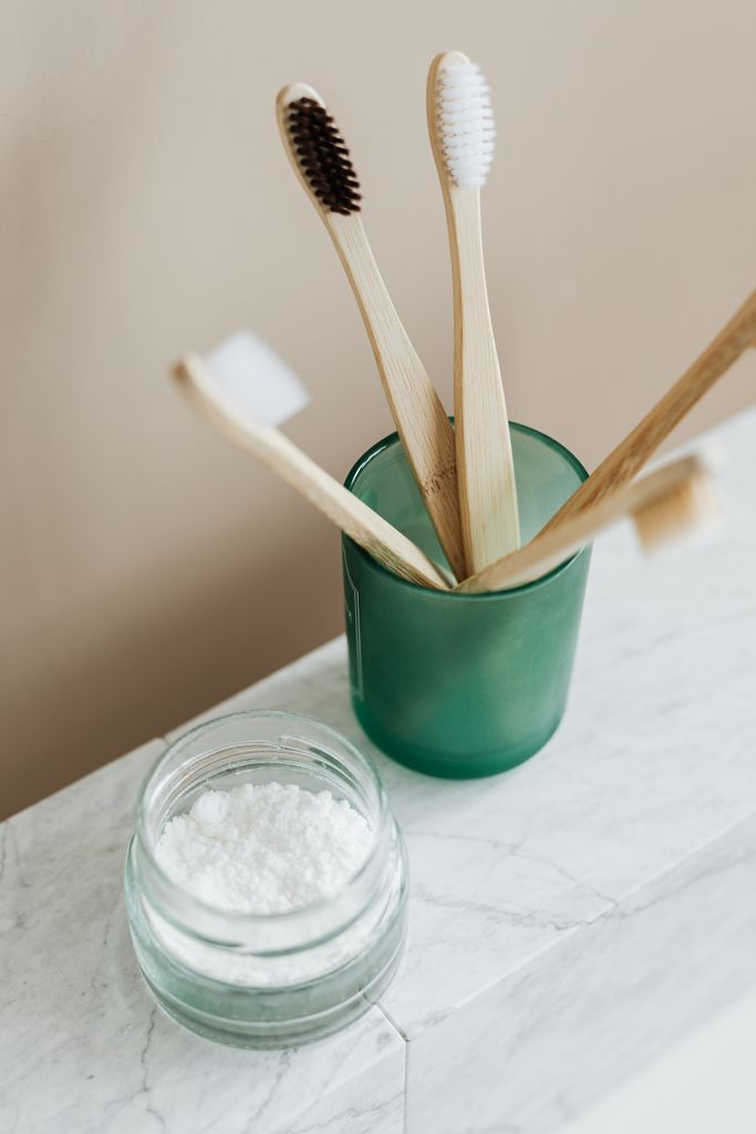From above of assorted bamboo eco friendly toothbrushes in translucent green holder near opened glass reusable jar with white toothpaste arranged on marble table against beige wall